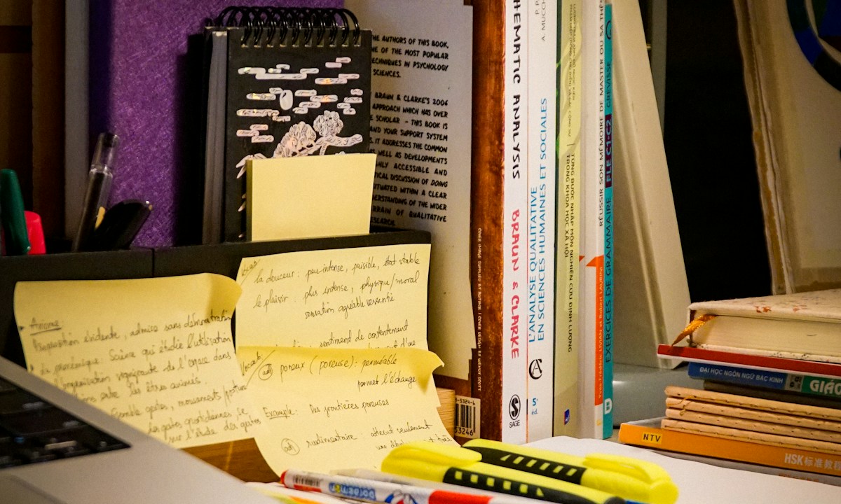 Boeken, handgeschreven notities en markeerstiften op een bureau naast een laptop.