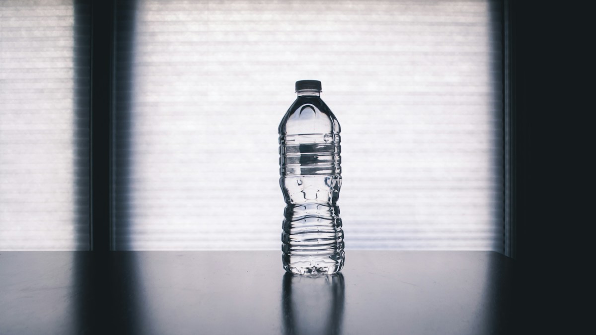 Een doorzichtige fles water met gesloten dop op een tafel voor een raam.